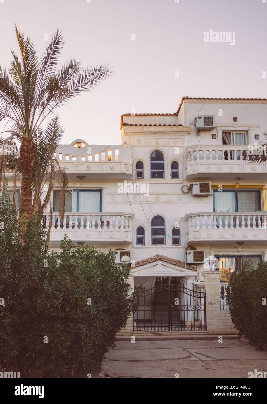 White house in Egypt with beautiful arched balconies and air conditioners. A palm tree and beautiful green bushes grow near the house. Paved backyard Stock Photo