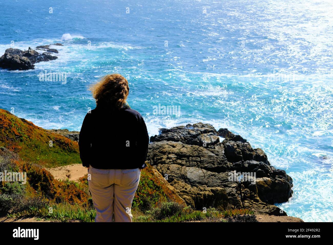 Middle-aged woman facing the ocean from above, overlooking rocks on the coast in Northern California; solitude, pensive outlook, facing the future. Stock Photo