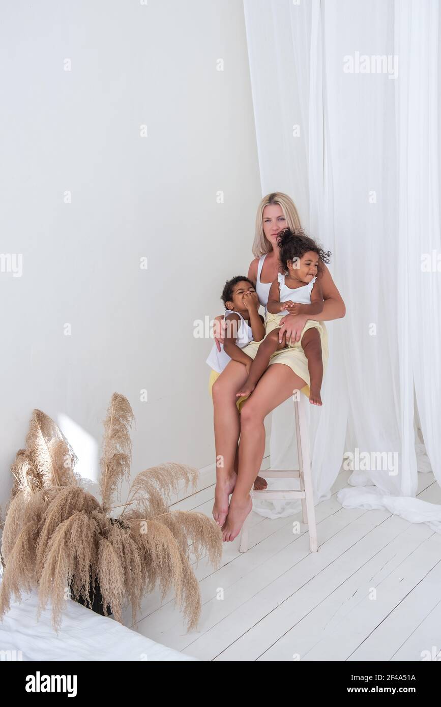 Young Caucasian mother sits on wooden chair, embrace her happy African American children. Daughter is sitting in the woman's arms, son hugging. Adopti Stock Photo