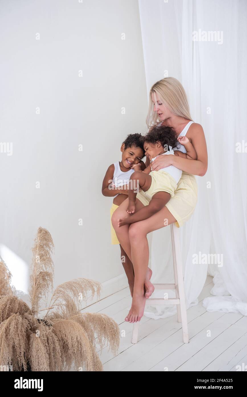 Young Caucasian mother sits on wooden chair, embrace her happy African American children. Daughter is sitting in the woman's arms, son hugging. Adopti Stock Photo