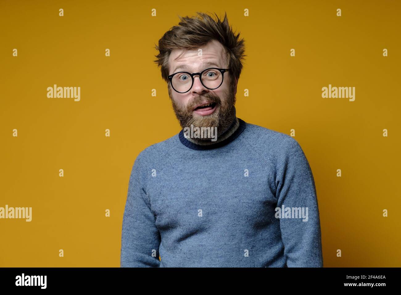 A strange shaggy man with glasses, with a stupid expression on his face, looks at the camera in bewilderment. Yellow background. Stock Photo