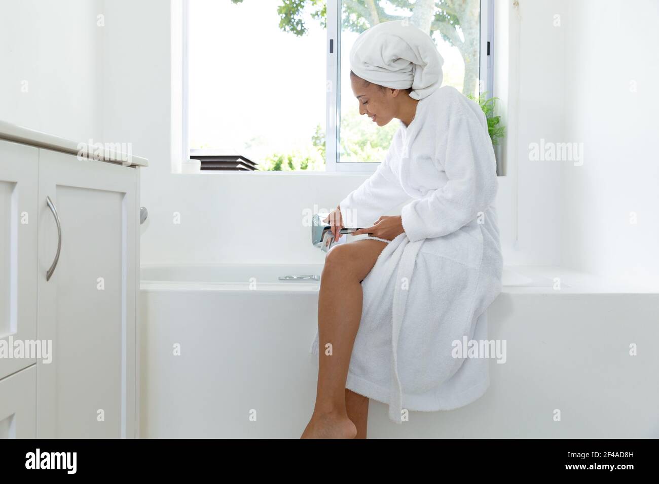 Mixed race woman wearing bathrobe sitting on bathtub edge in bathroom Stock Photo