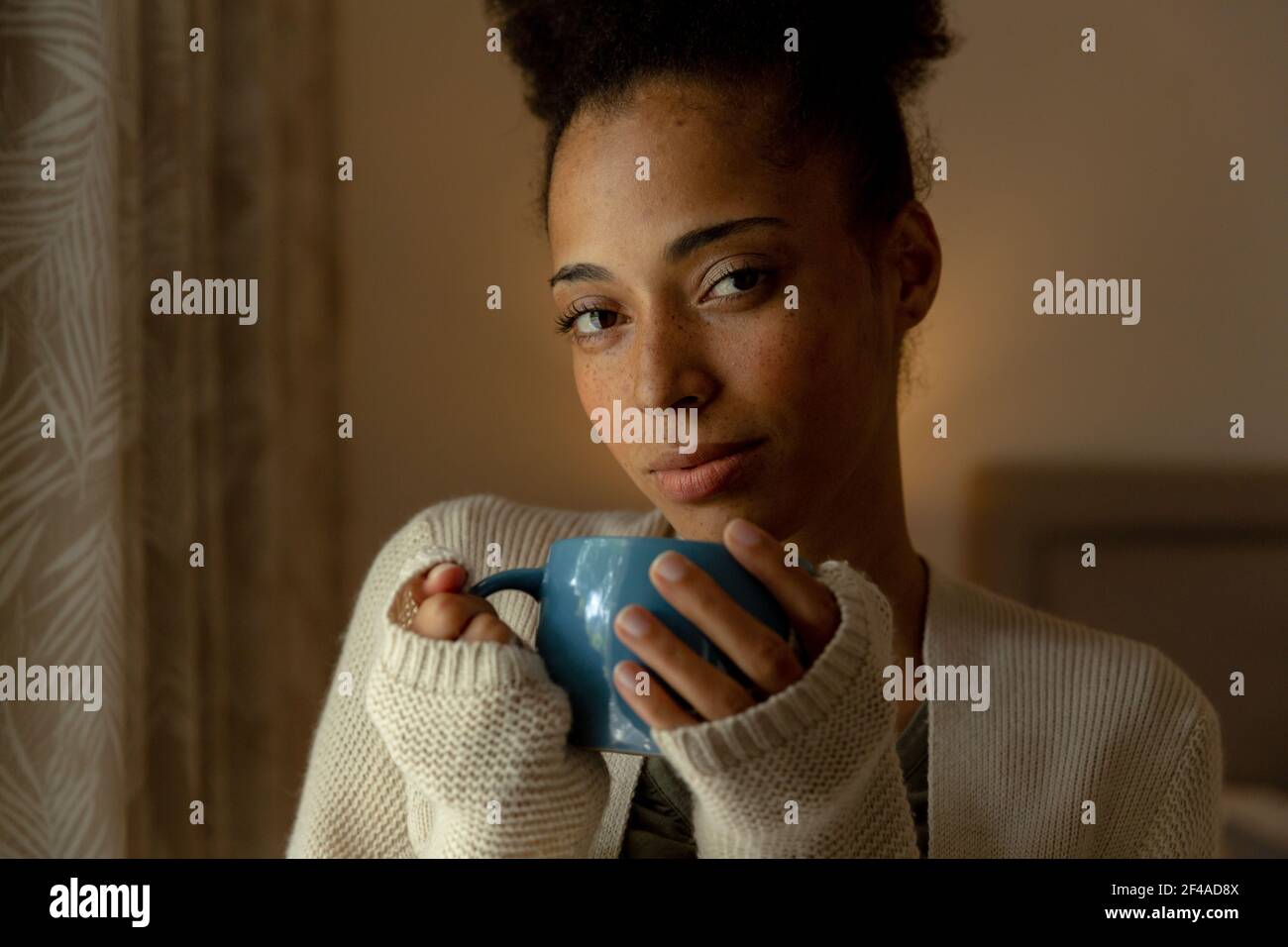 Mixed race woman standing by a window and drinking cup of coffee at home Stock Photo
