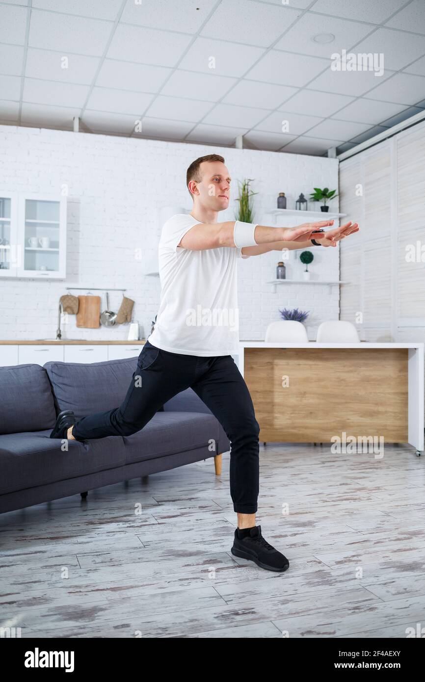 Handsome sporty man in a T-shirt doing squats at home in his apartment. Fitness at home. Healthy lifestyle Stock Photo