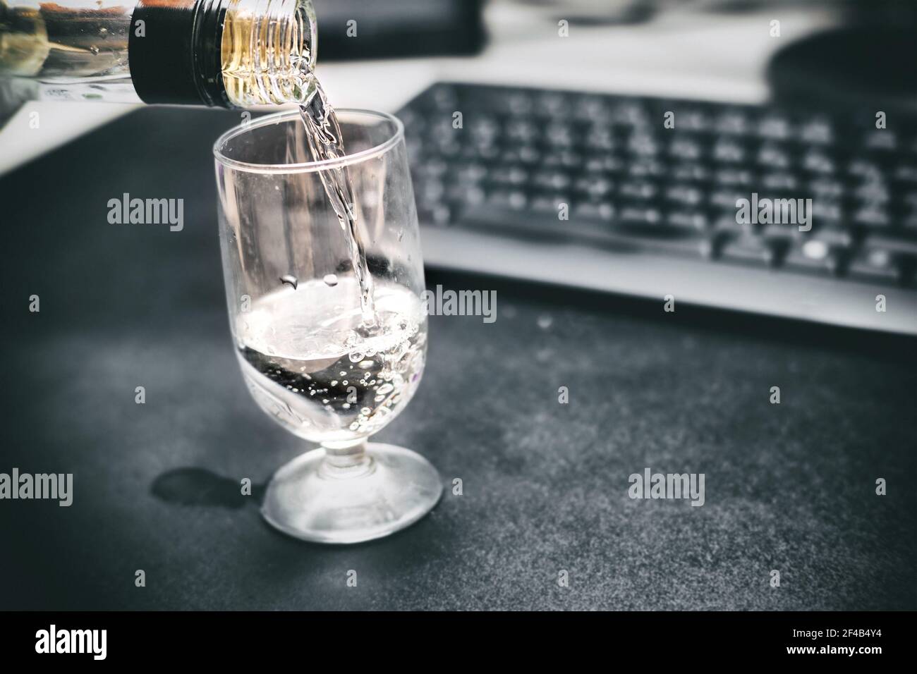 Drunk at work. Pouring a glass of vodka. Defocused office desk and keyboard to show compromised vision and brain function. Concept for coping with wor Stock Photo