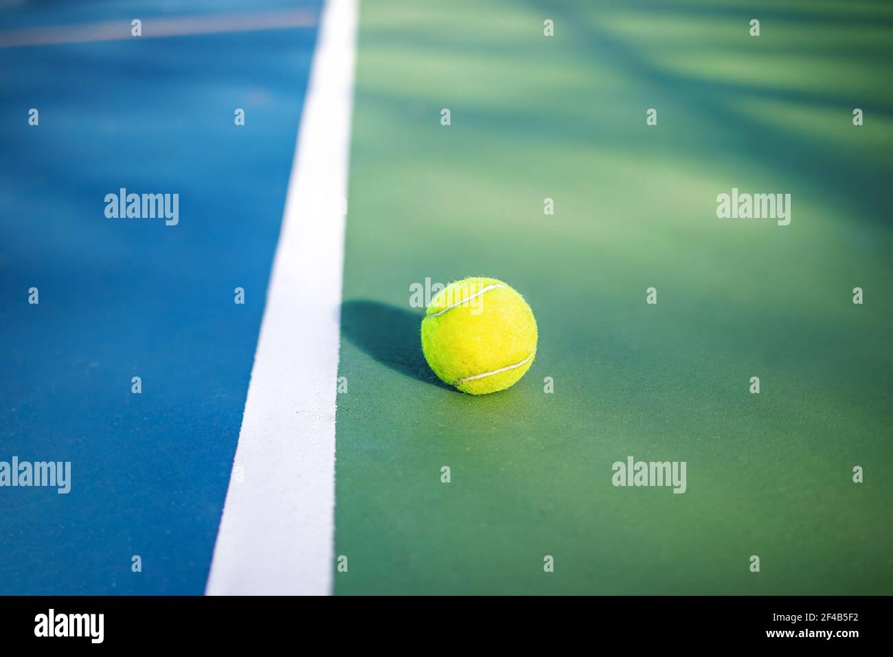 Yellow tennis ball next to sideline in outdoor tennis court, closeup. Defocused and abstract blue and green rubberized ground surface for shock absorp Stock Photo