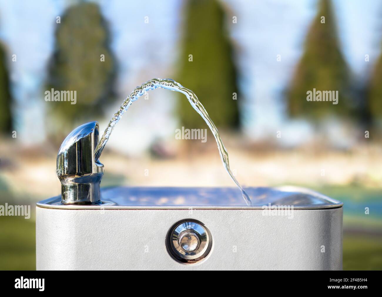 Drinking water fountain in park on a sunny day, no person. Close up of flowing water from the tab in an arch. Bright defocused park. Stock Photo