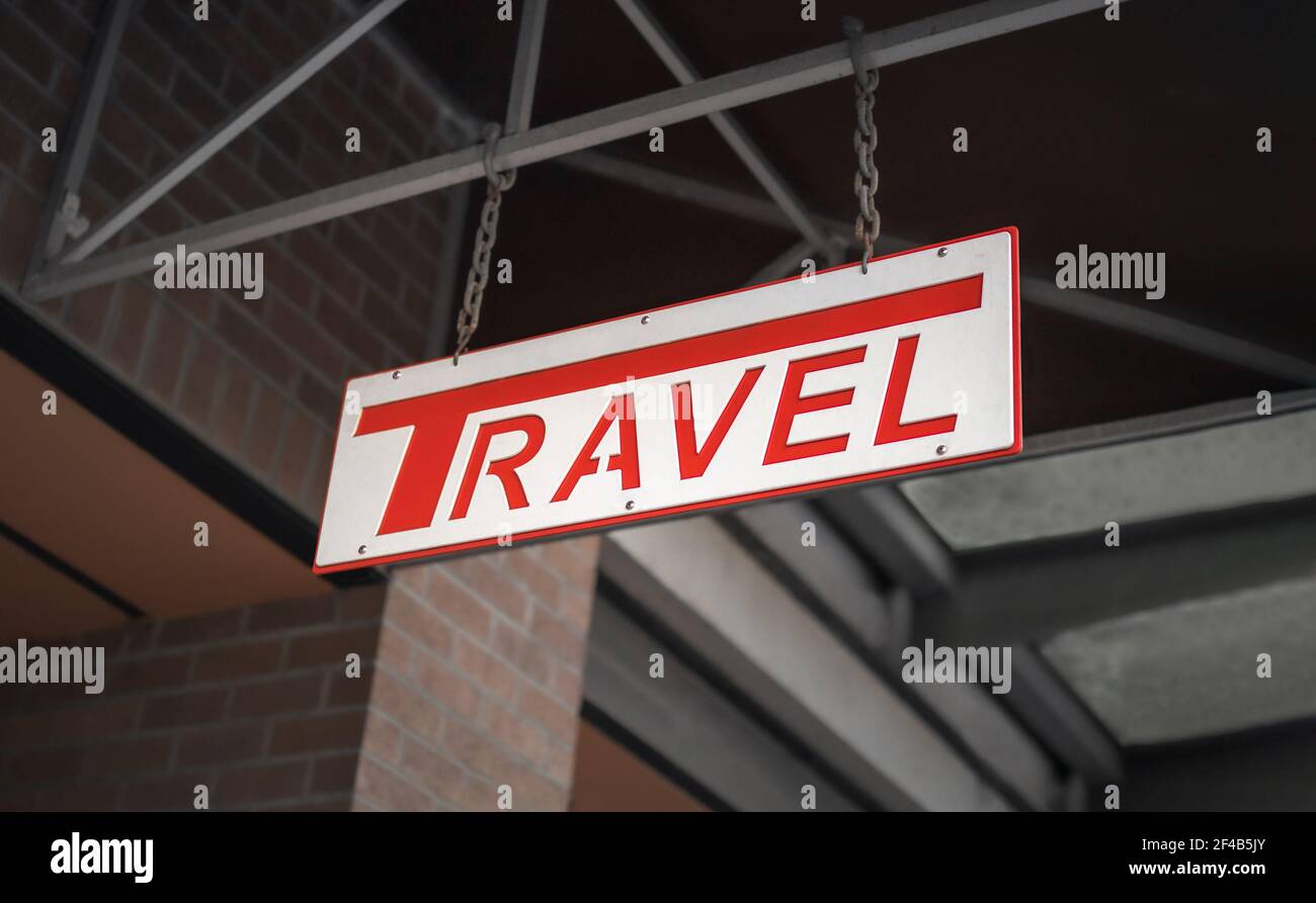 Metal travel sign hanging of a building outside. White store sign with debossed red 'Travel' text. Tourism travel agency signage. Selective focus. Stock Photo