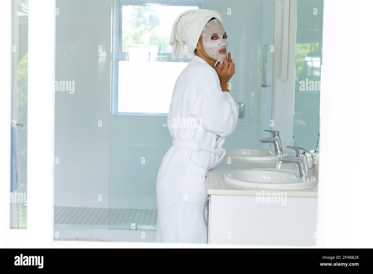 Mixed race woman wearing bathrobe and cleansing face mask in bathroom Stock Photo