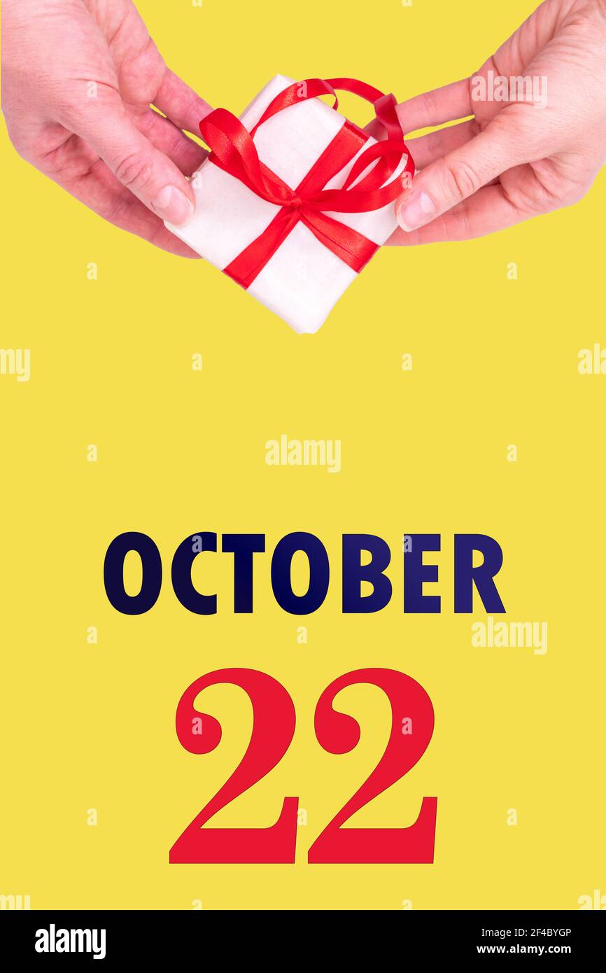 October 22nd. Festive Vertical Calendar With Hands Holding White Gift Box With Red Ribbon And Calendar Date 22 October On Illuminating Yellow Backgrou Stock Photo