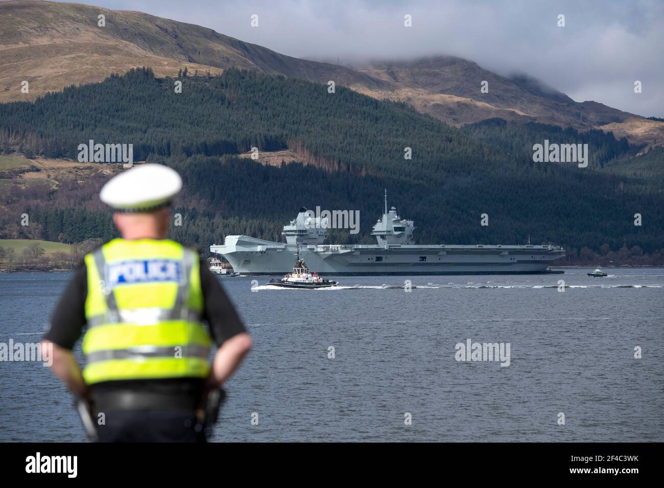 Cove, Argyle and Bute, Scotland, UK. 20th Mar, 2021. PICTURED: A police officer watches on as HMS Queen Elizabeth sails into the Firth of Clyde. People turn out to watch HMS Queen Elizabeth set sail after almost a week being berthed in Loch Long at Glenmallan where it took on fuel, munitions and other supplies, ahead of naval exercises which are part of the UK Carrier Strike Group 2021. Credit: Colin Fisher/Alamy Live News Stock Photo