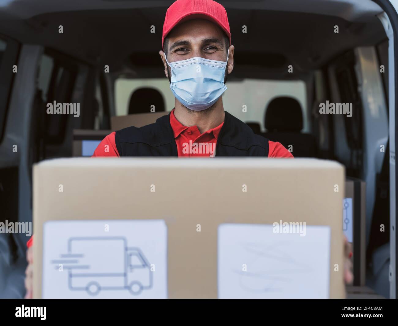 Young delivery man carrying cardboard box while wearing face mask to avoid corona virus spread - People working with fast deliver during corona virus Stock Photo