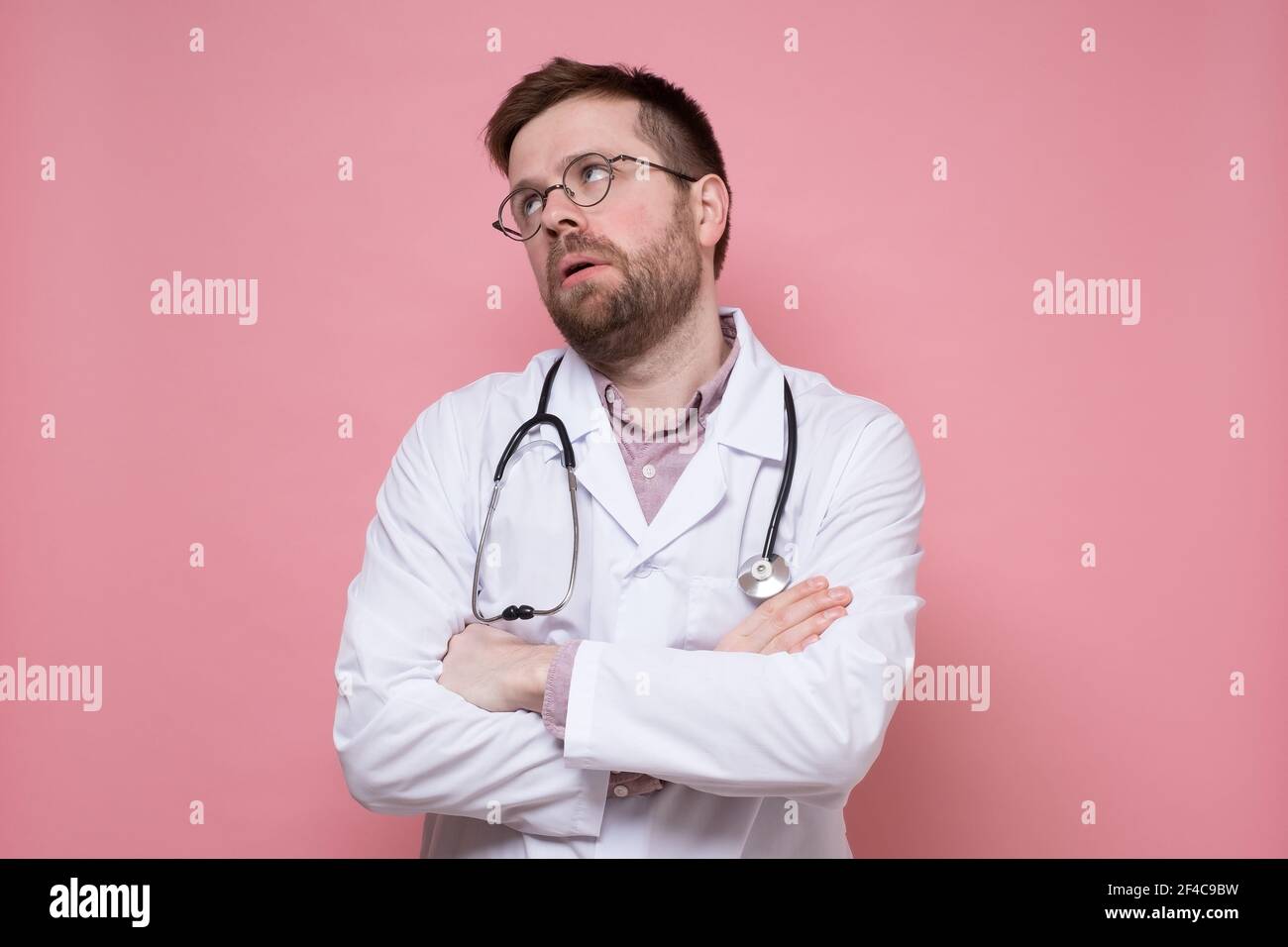 Tired male doctor in a white coat, with a stethoscope around neck, crossed arms and looked up apathetically. Pink background. Stock Photo