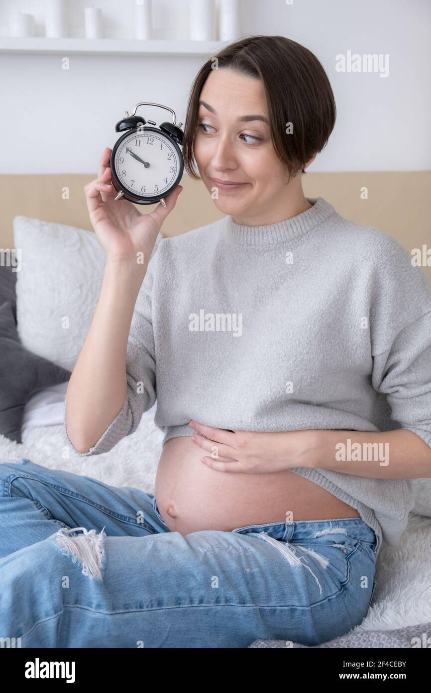 Pregnant young adult woman with alarm clock on bed at home, giving birth soon concept Stock Photo