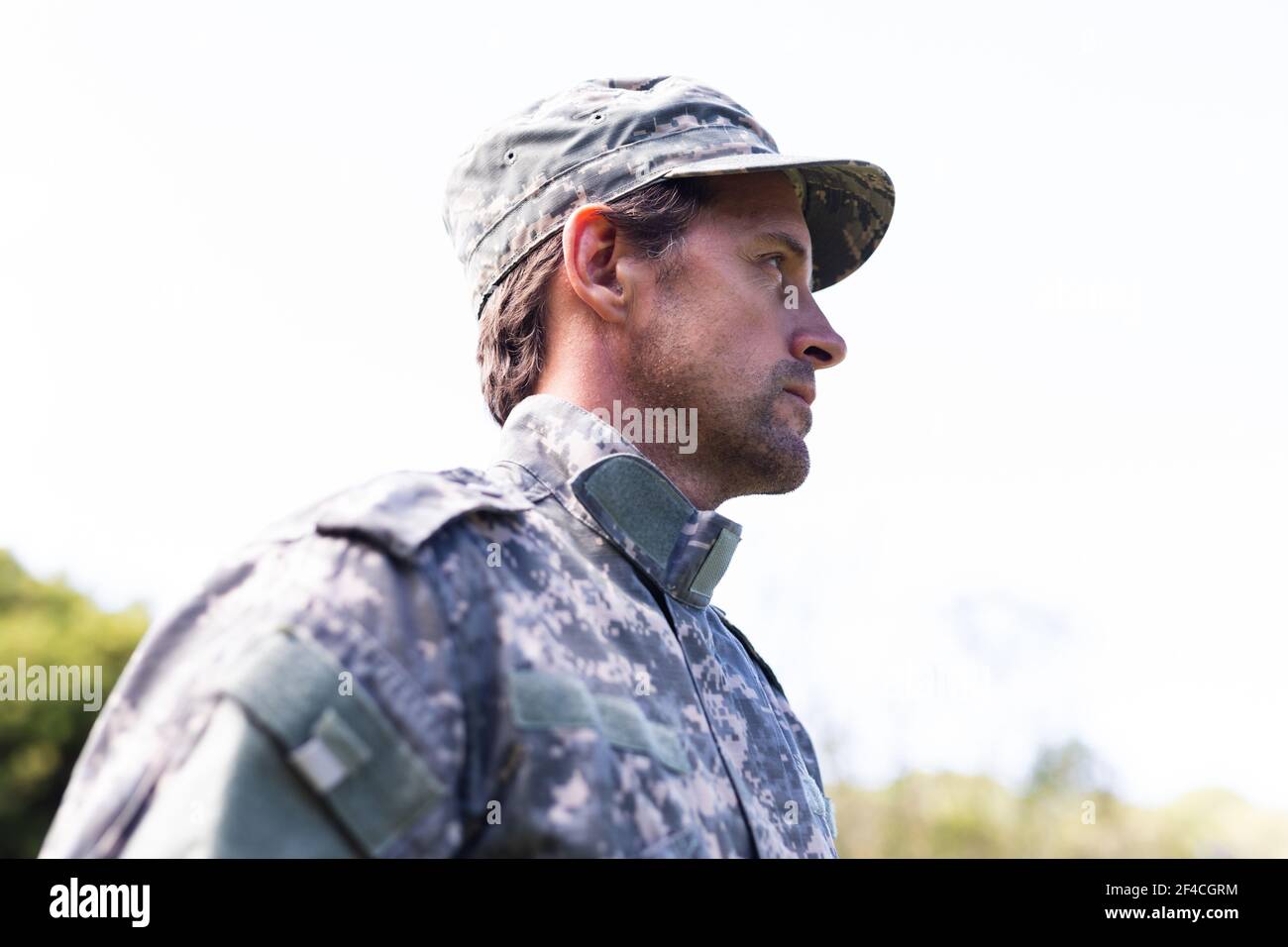 Caucasian male soldier wearing camo fatigues and cap standing outdoors looking away Stock Photo