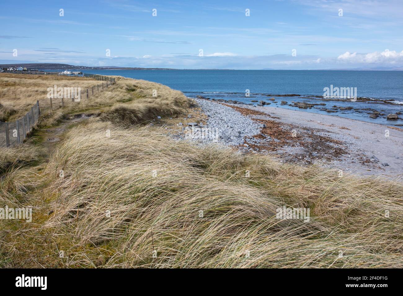 Landscape with grey pebble beach, beige dry grass, blue ocean and sunny, blue sky on Inisheer island, the smallest of Aran islands. Stock Photo