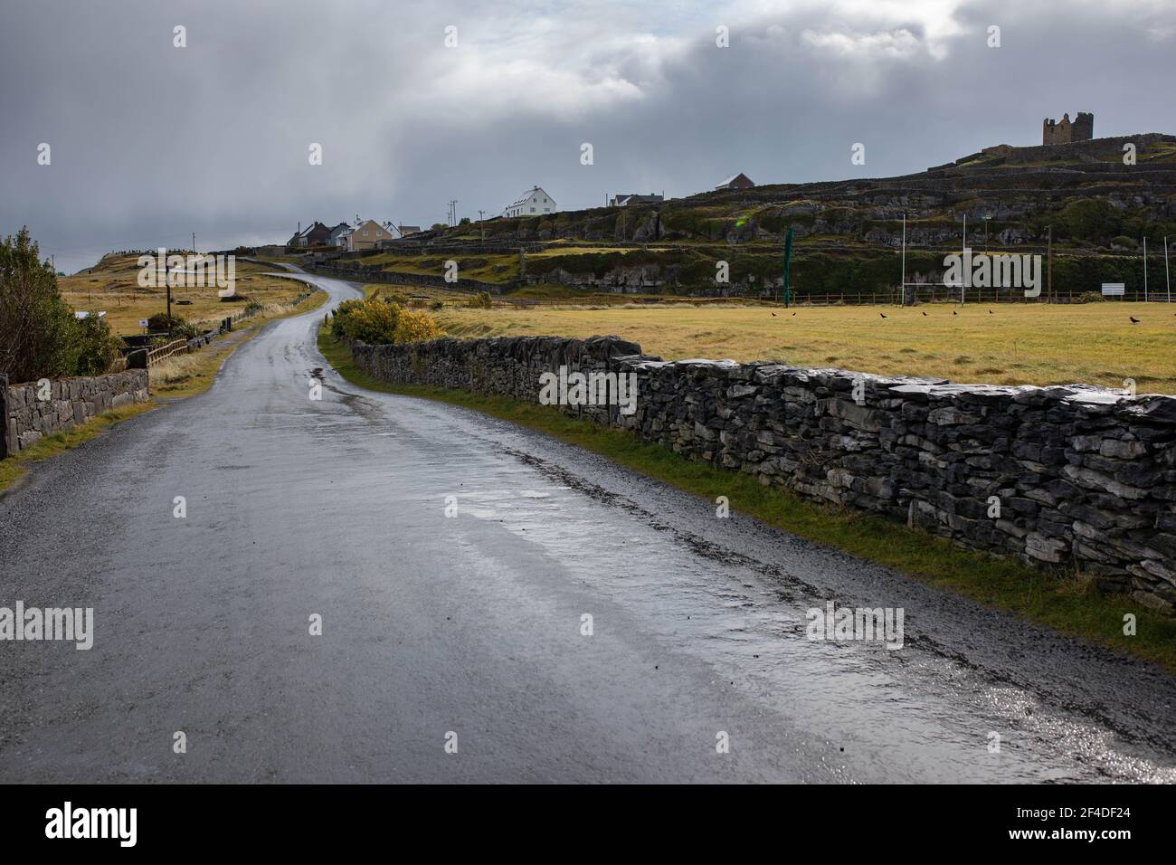 Landscape with grey, wet road going up hill, stonewall, mustard color grass, grey sky, castle ruins on the hill on an overcast autumn day on Inisheer Stock Photo