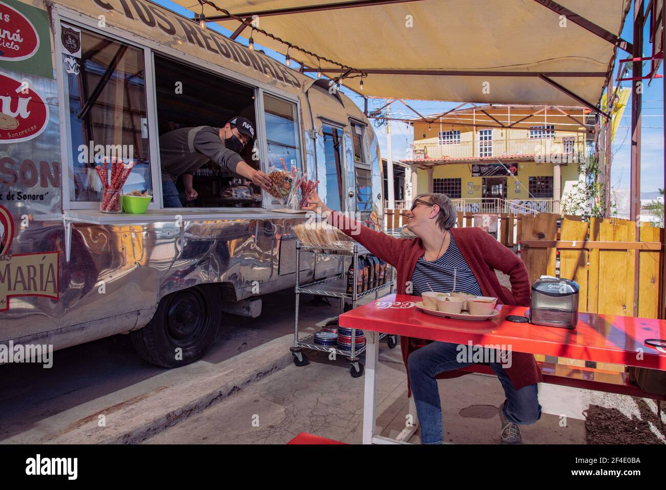 Woman sitting outside at red table reaches for candy held by employee  in a food truck in Santa Ana, Sonora, Mexico. Stock Photo