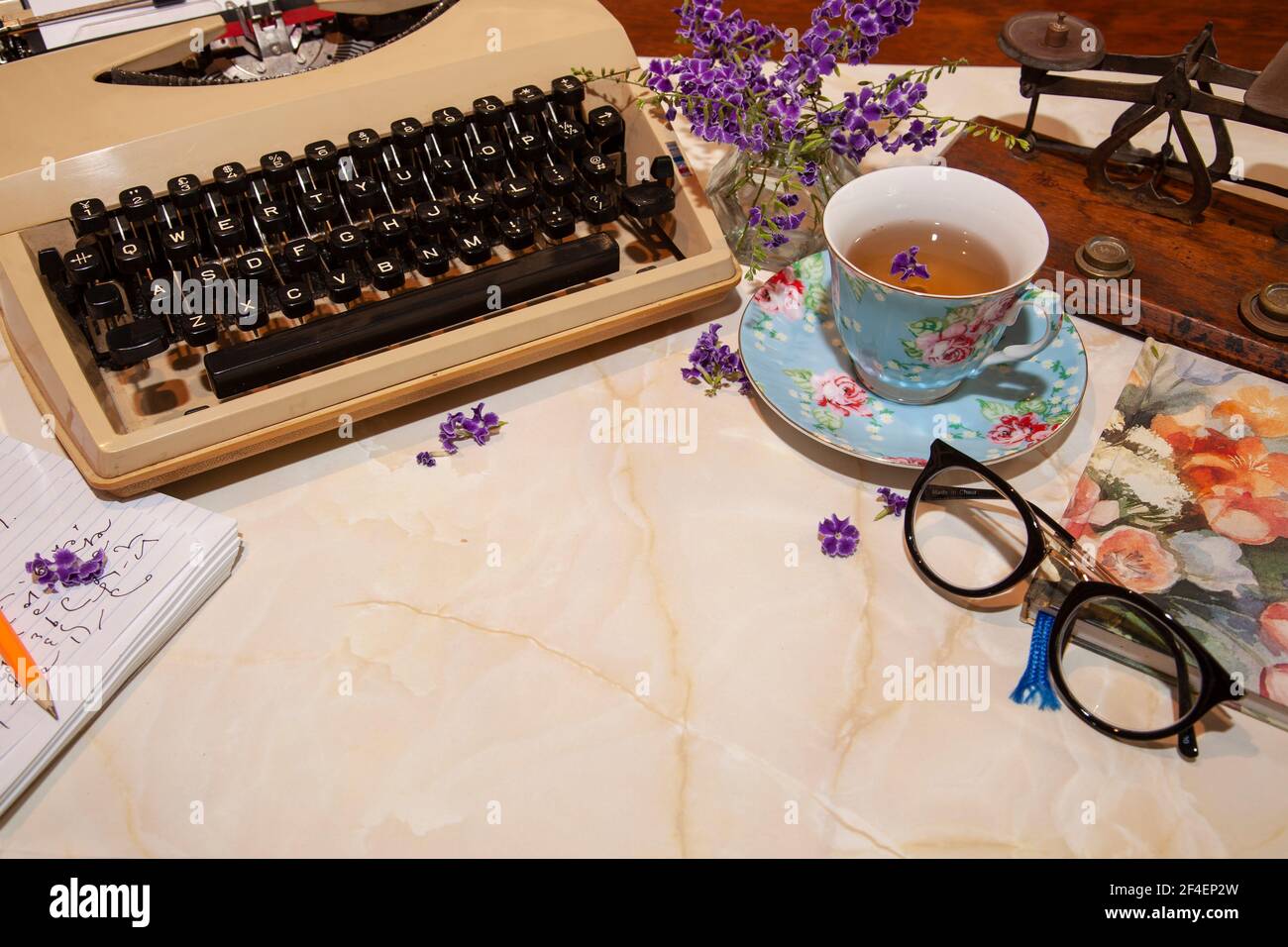 An old fashioned retro manual typewriter on a desk with cup, notepad, spectacles, letter scales and flowers with copy space. Stock Photo