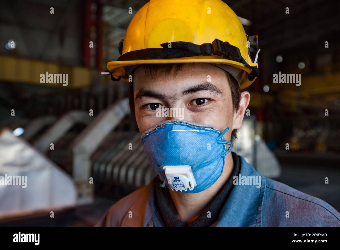 Aluminum electrolysis plant. Young Asian worker portrait. Slag handling crane operator. In yellow hardhat and blue respirator. Electrolysis baths on b Stock Photo