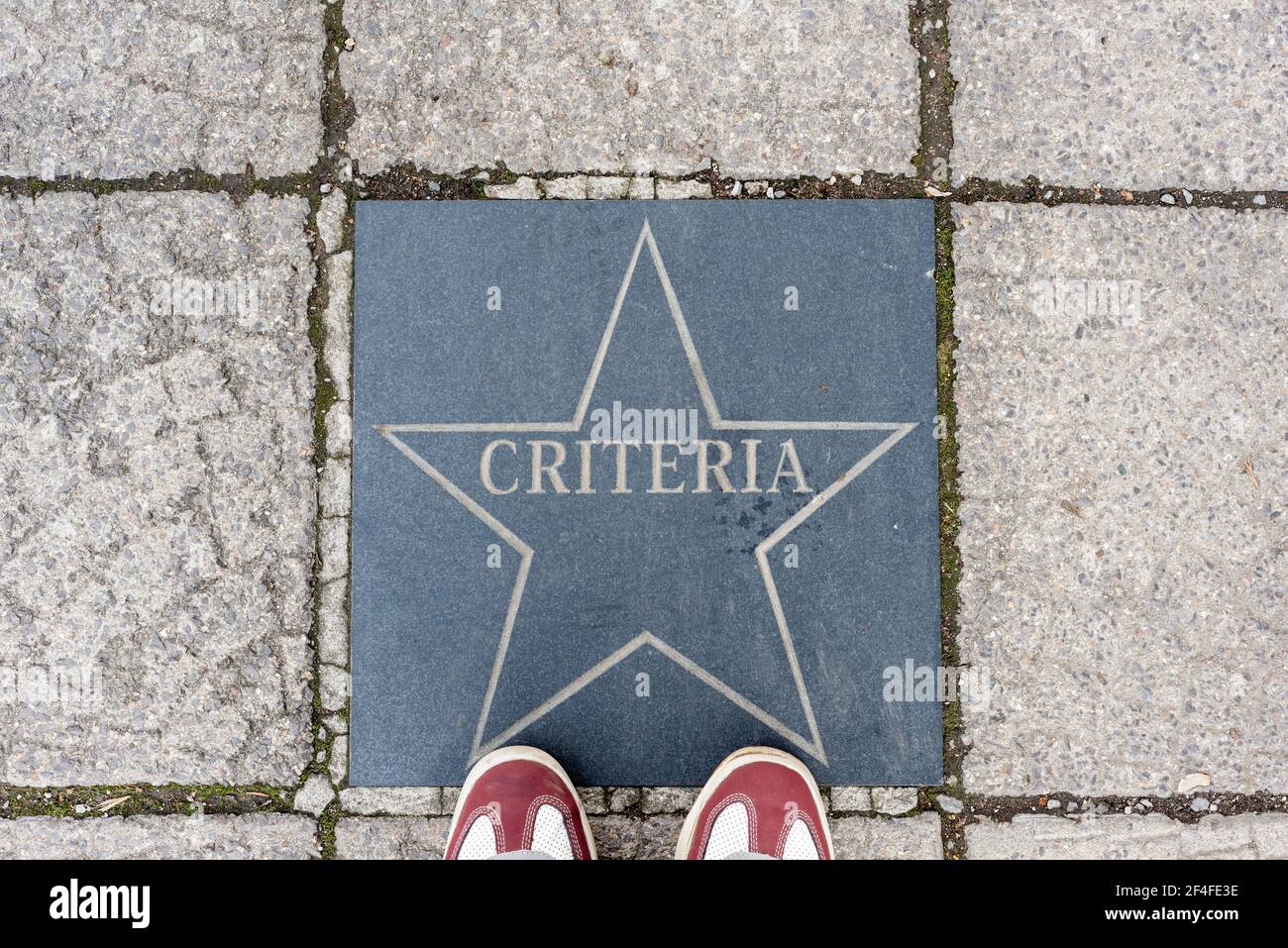 Criteria text in star shaped sign on sidewalk or pavement and sneakers partly visible, top view. Concept. Stock Photo