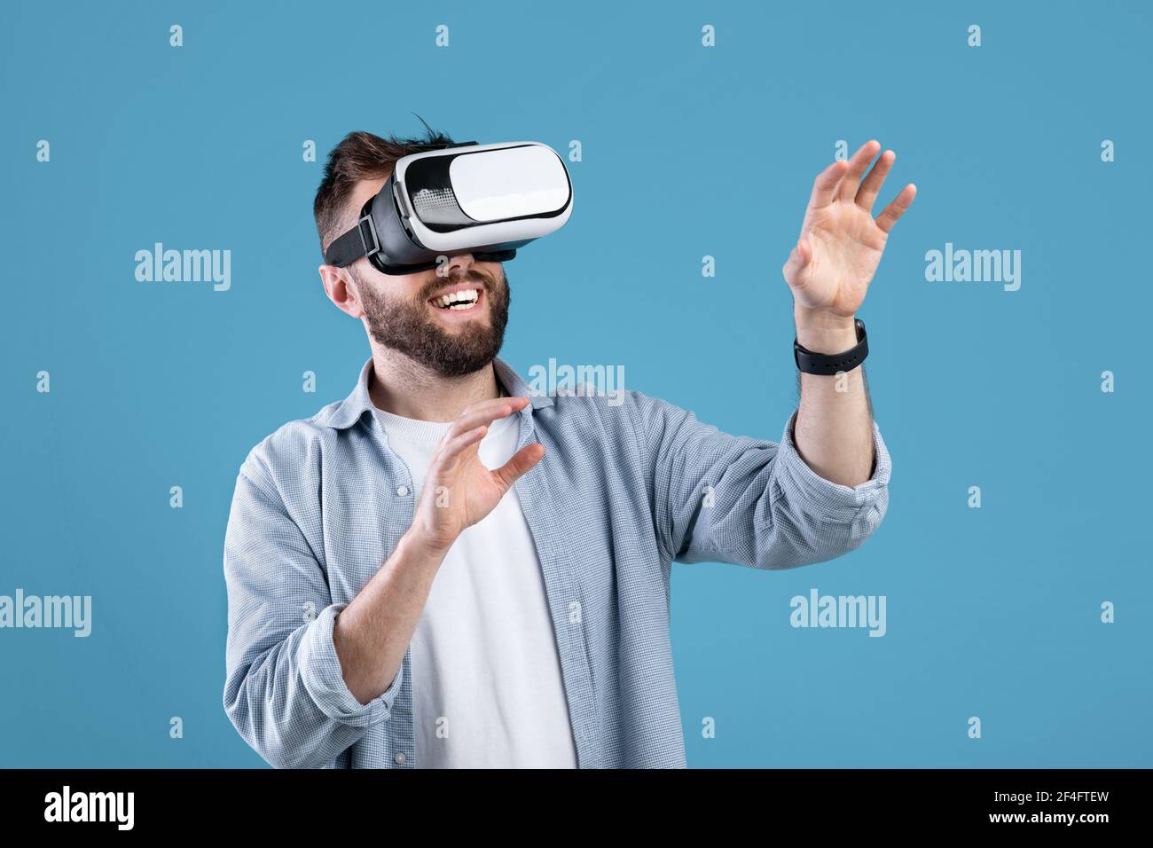 Happy bearded young man using VR headset to explore augmented reality on blue studio background Stock Photo