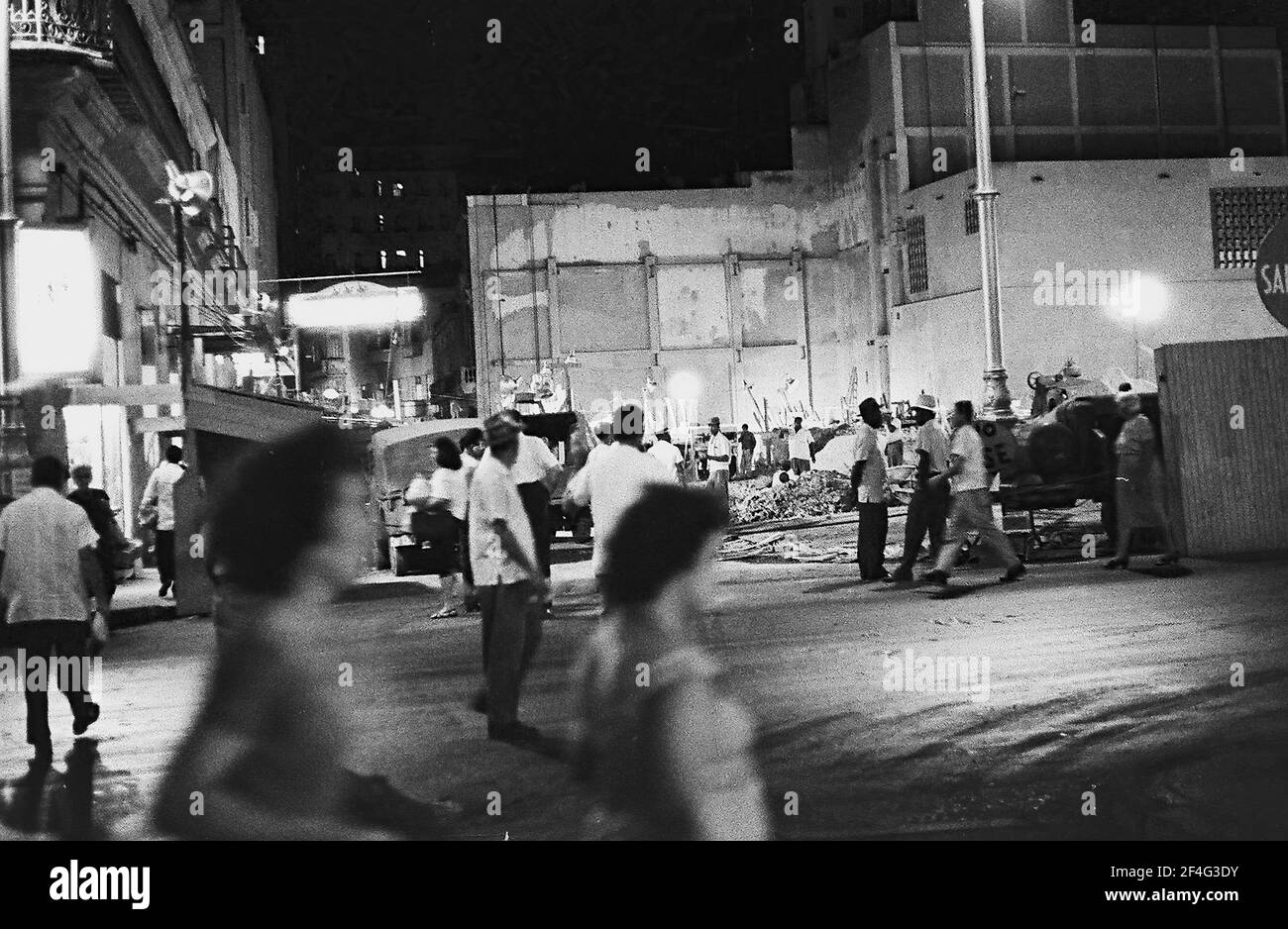 Angled night-shot of people walking in a well-lit urban area, with a rubble-filled lot in the background, Havana, Cuba, 1964. From the Deena Stryker photographs collection. () Stock Photo
