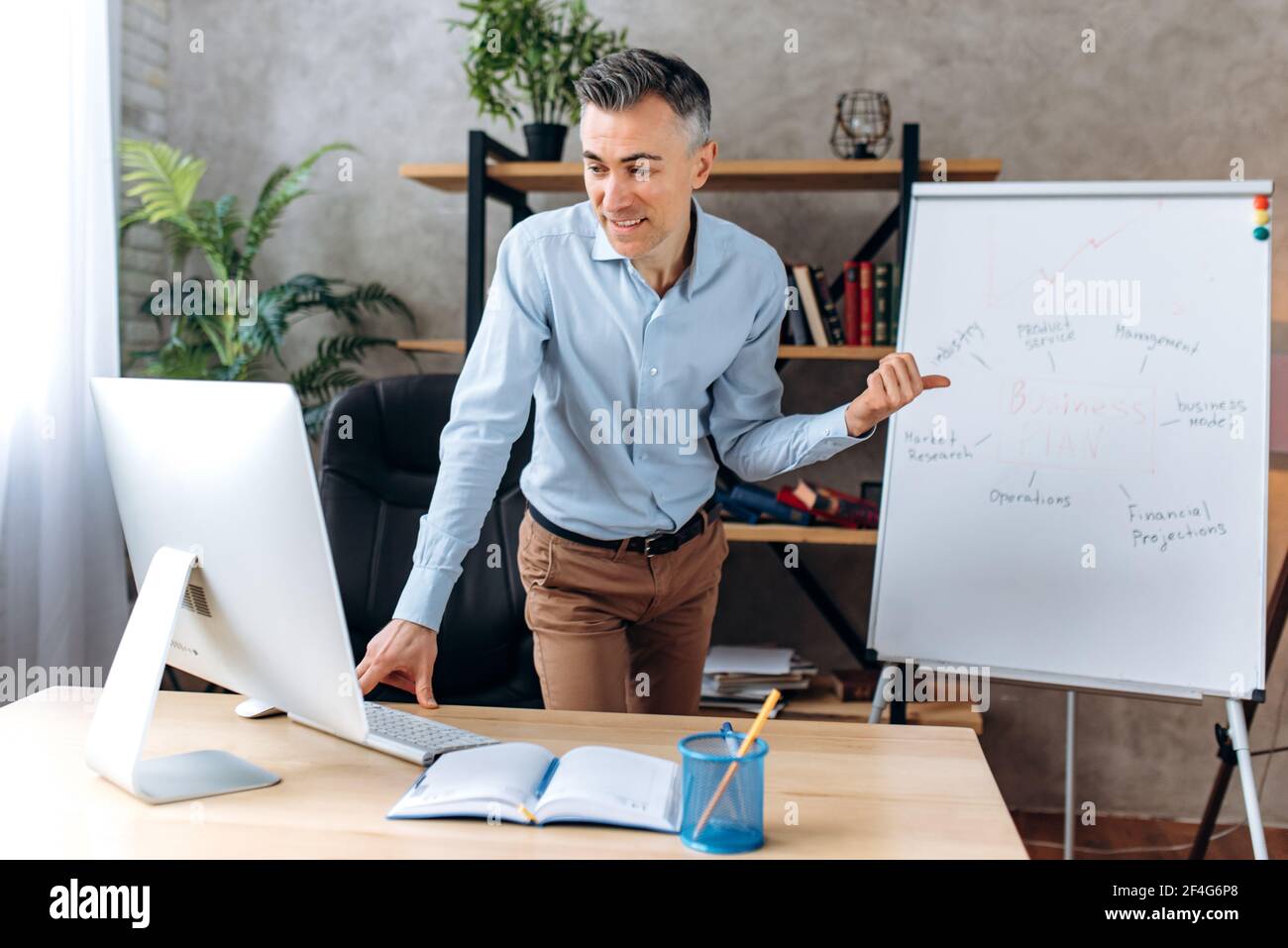 A successful confident adult businessman, the leader of the company conducts online business meeting for colleagues in office, gestures with hand, explain business strategy, shows on flipchart, smiles Stock Photo