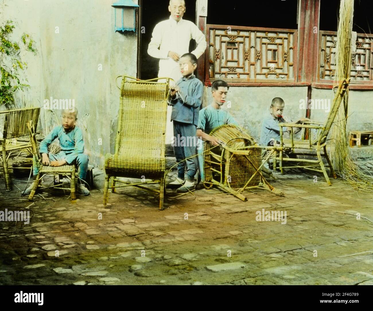 A man stands in a doorway, facing the camera, while several boys sit and stand while caning chairs in a courtyard at a School for the Blind, Beijing, China, 1918. From the Sidney D. Gamble photographs collection. () Stock Photo