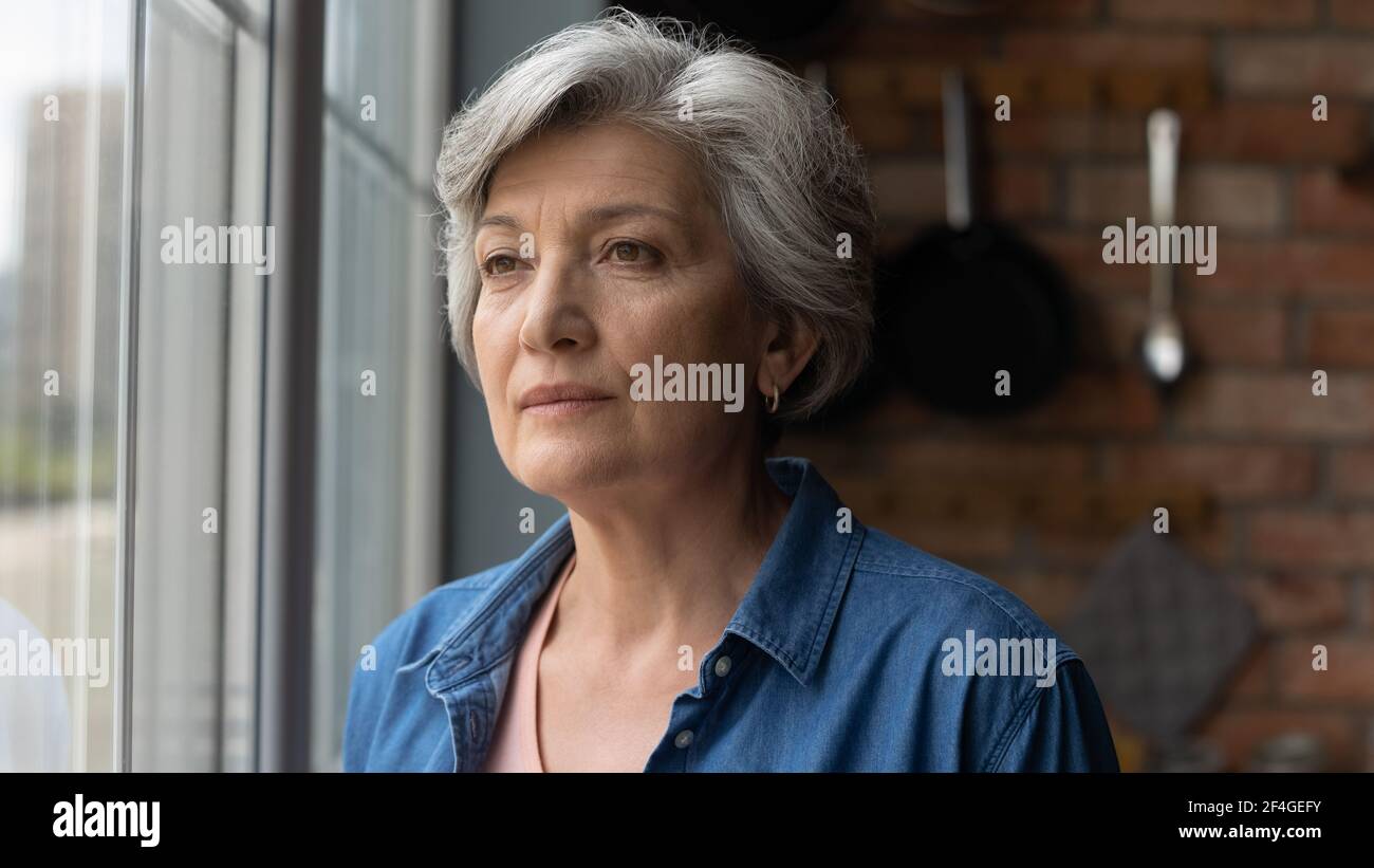 Worried depressed old latin lady stand by window look away Stock Photo