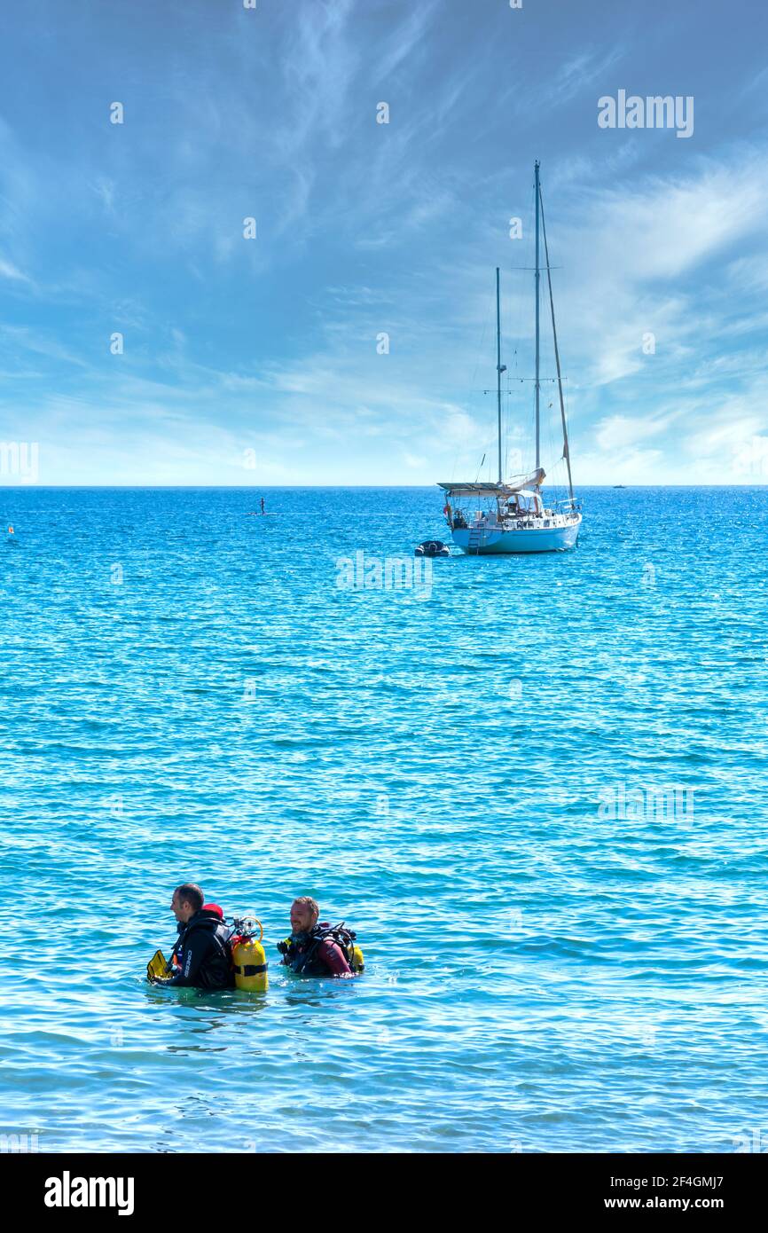 La Herradura, Spain; October-17, 2020: Two men dressed in wetsuits coming out of the sea with a nice sailboat in the background Stock Photo