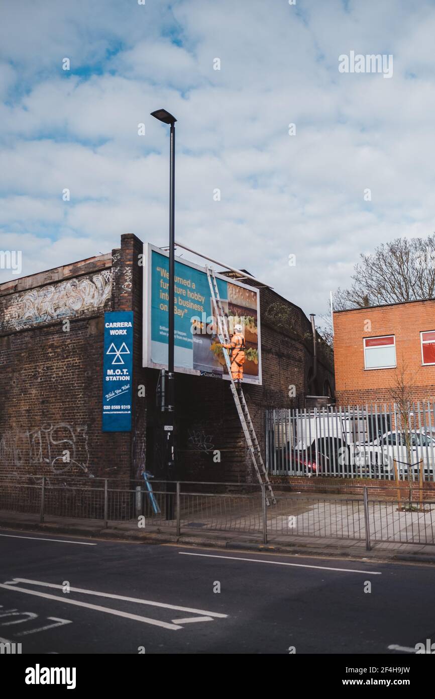 Brentford, West London | UK -  2021.03.21: Worker prepares billboard to installing new advertisement. Industrial climber working on a ladder - placing Stock Photo