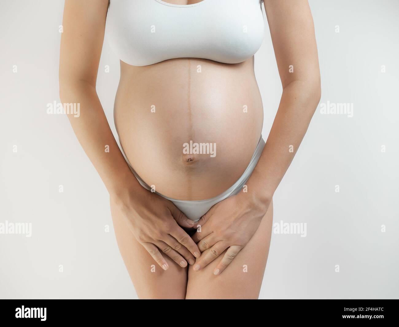 A woman in the third trimester of pregnancy. A pregnant young woman with dark hair stands holds hands in the lower belly against a white background. T Stock Photo