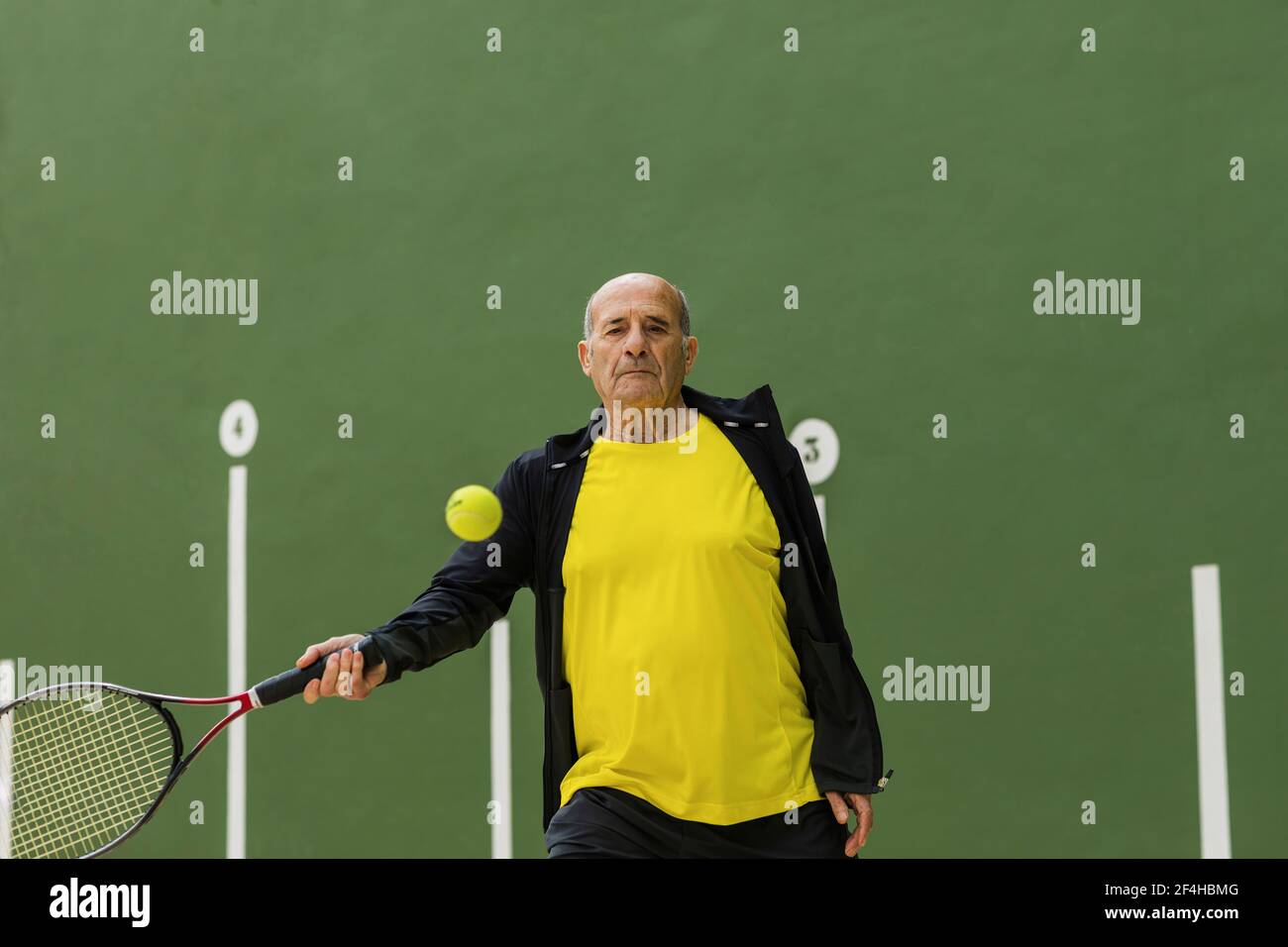 Elderly male athlete hitting ball with racket while playing tennis against  green wall in gym Stock Photo - Alamy