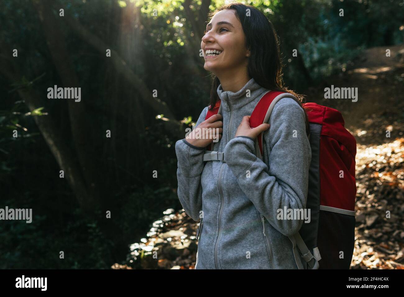 Happy optimistic young female hiker in activewear with backpack enjoying journey in green forest in sunny day Stock Photo