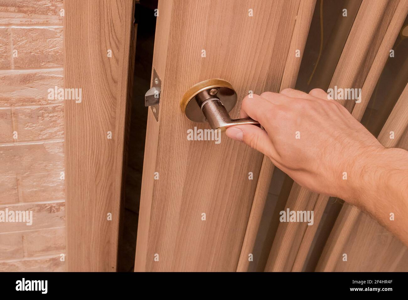A man's hand holding an iron door handle opens or closes a wooden door indoors. Stock Photo