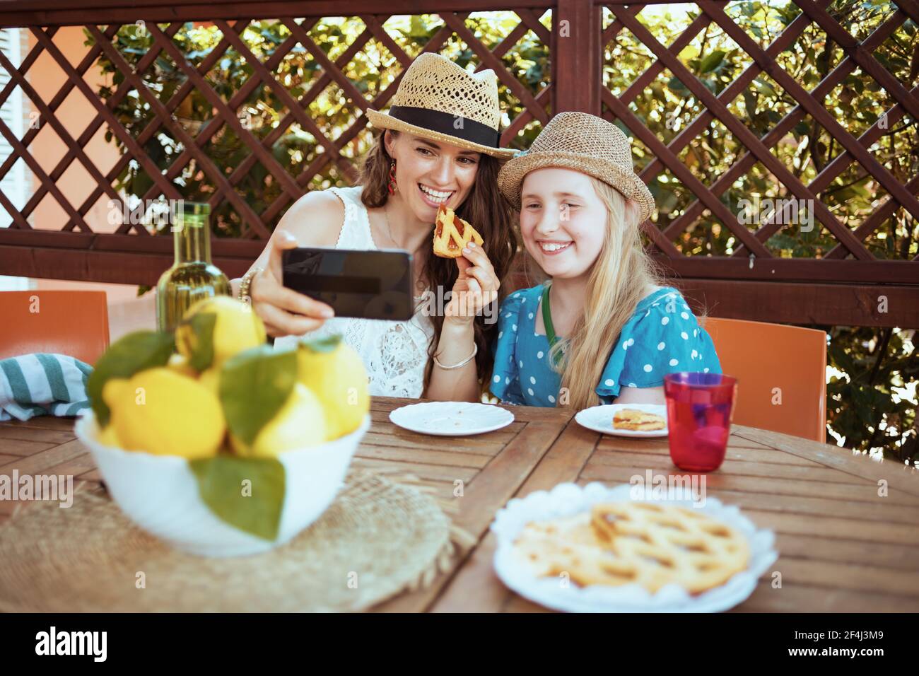 smiling stylish mother and daughter with crostata and plate of local farm lemons having video meeting on a smartphone and having lunch in the patio. Stock Photo