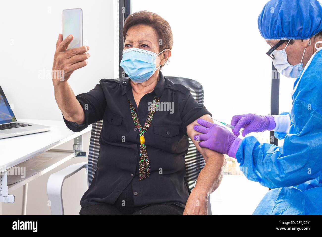 Elderly adult woman taking a selfie with her cell phone while receiving the coronavirus vaccine in a hospital from a doctor during the pandemic. Techn Stock Photo