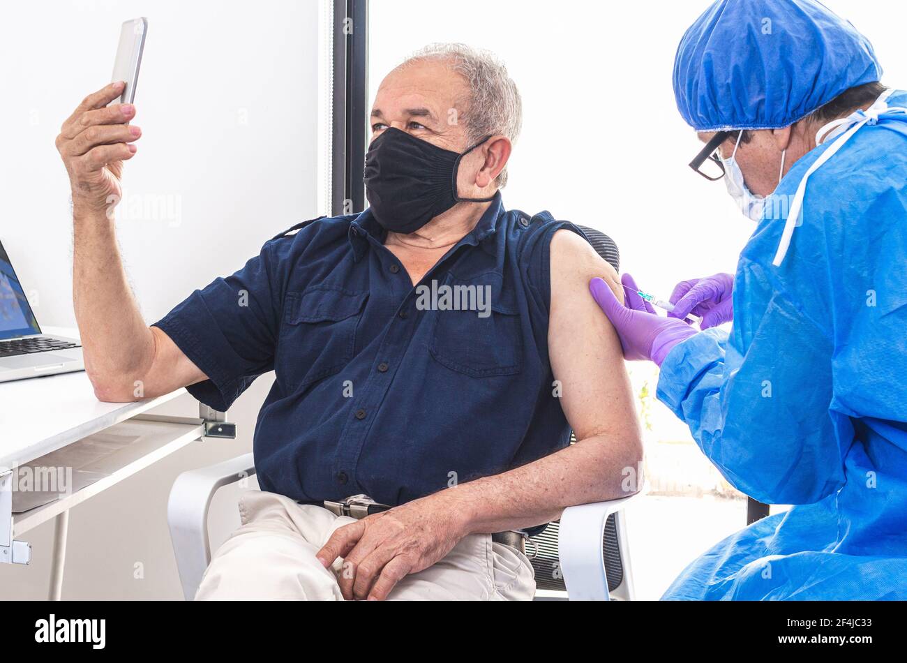 Adult, senior man taking a selfie with his cell phone while receiving the coronavirus vaccine in a hospital from a doctor during the pandemic. Technol Stock Photo