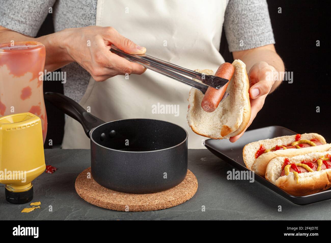 A caucasian woman is taking a fresh boiled hot dog sausage from a pot  using metal tong in order to place it into a hot dog bun. Hot pot is on a cork Stock Photo