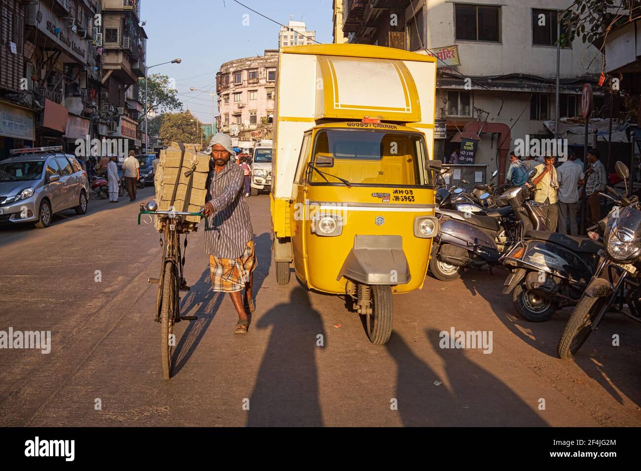 A small-time courier / porter pushes his loaded bicycle past a small three-wheeler truck; Mumbai, India Stock Photo
