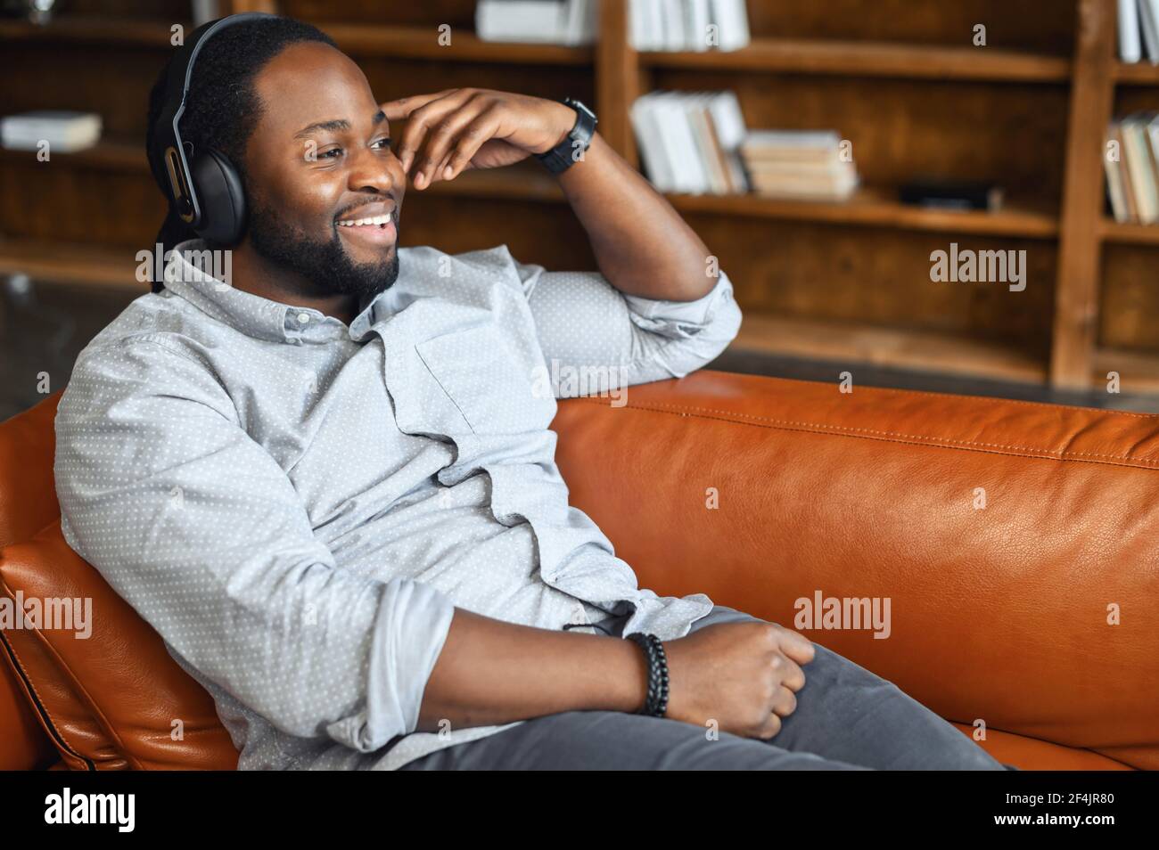 Relaxed and positive young African American man lying down on the couch in living room holding head and chilling enjoying listening to favorite music in wireless headphones spending his leisure time S...