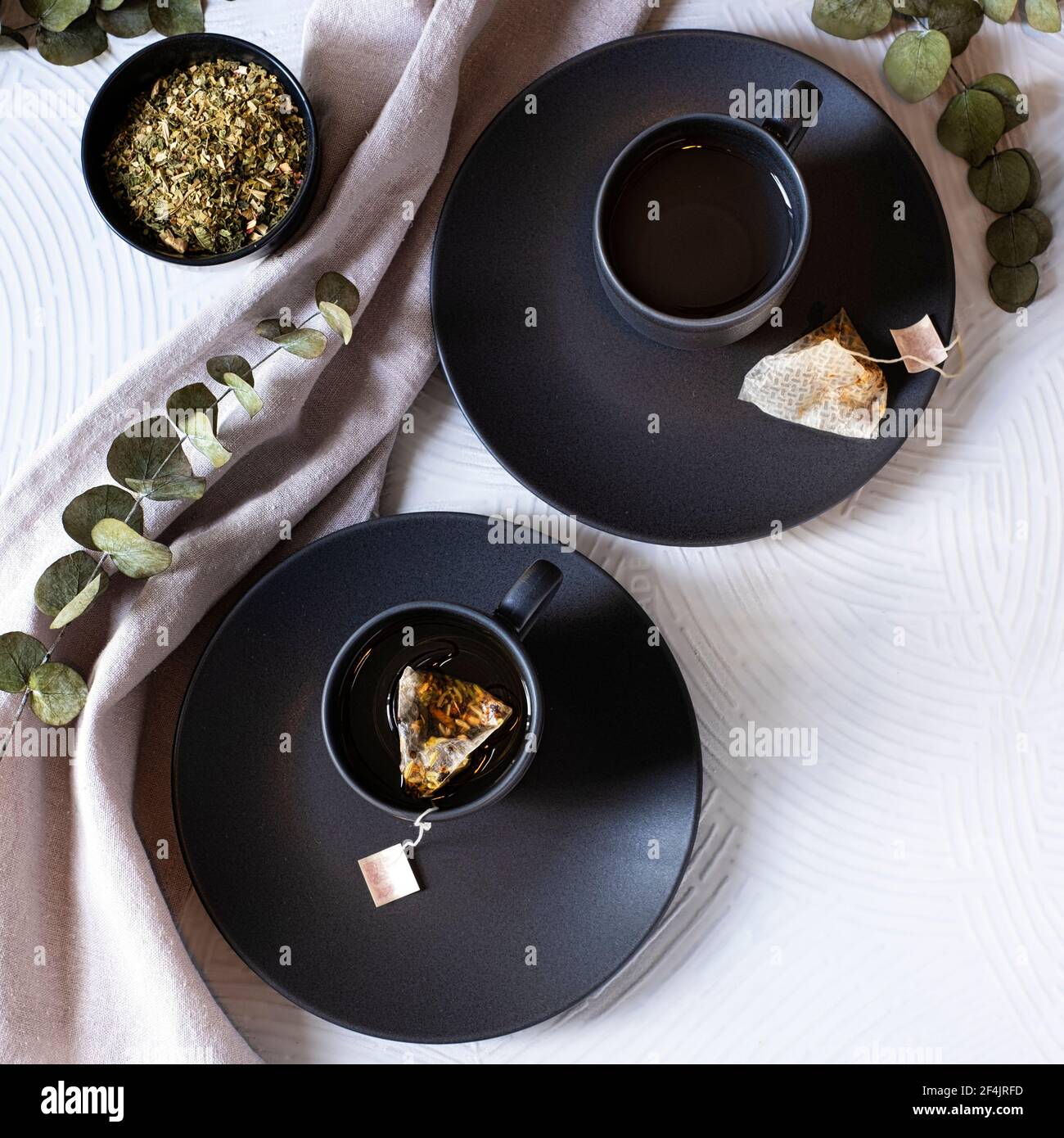 Two identical black tea cups on clay surrounded by eucalyptus branches on a white background Stock Photo