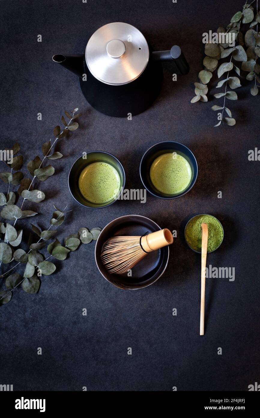 Two identical black tea cups on clay surrounded by eucalyptus branches, a bowl of macha tea with bamboo spoon and bamboo broom on a black background Stock Photo