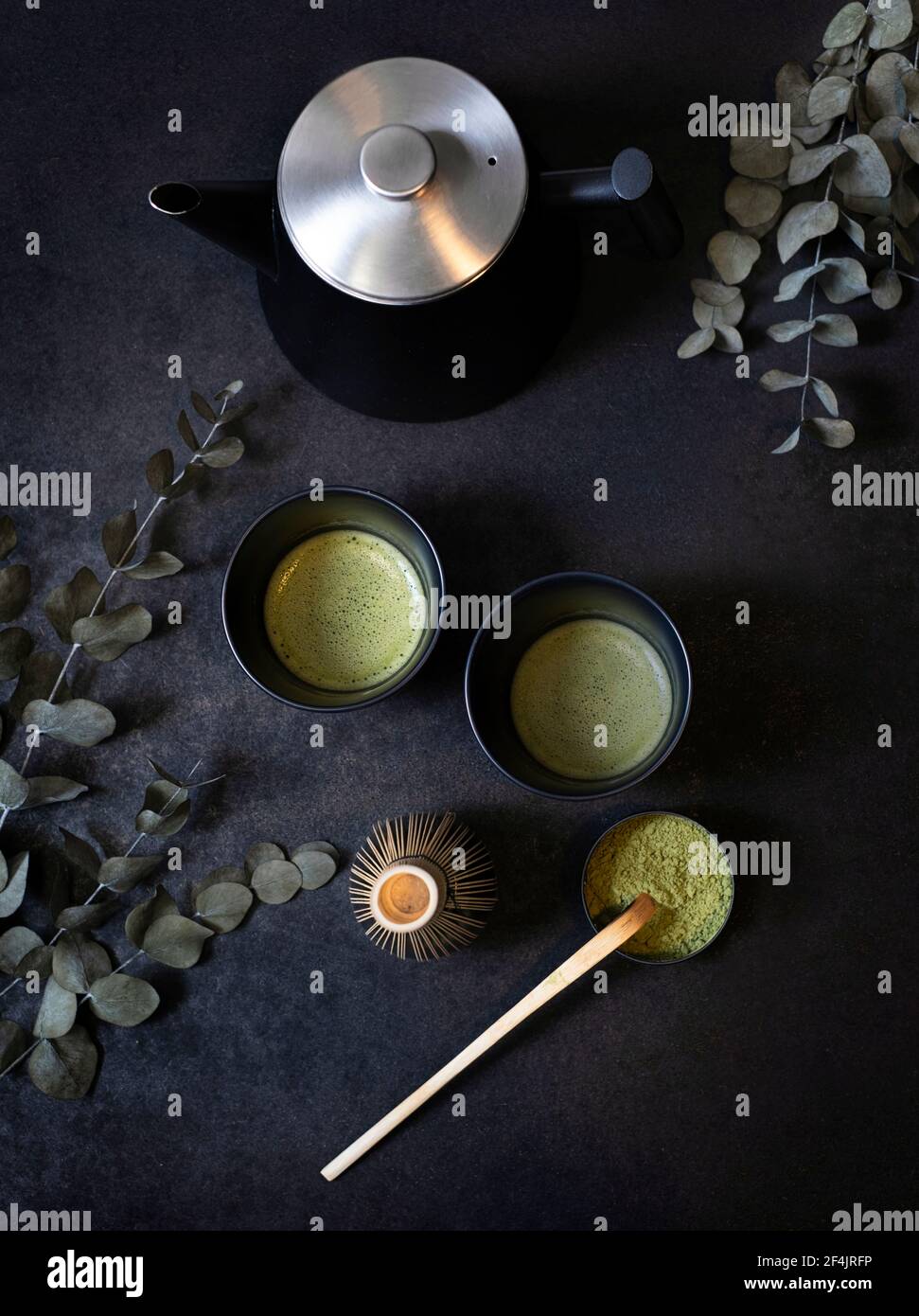Two identical black tea cups on clay surrounded by eucalyptus branches, a bowl of macha tea with bamboo spoon and bamboo broom on a black background Stock Photo