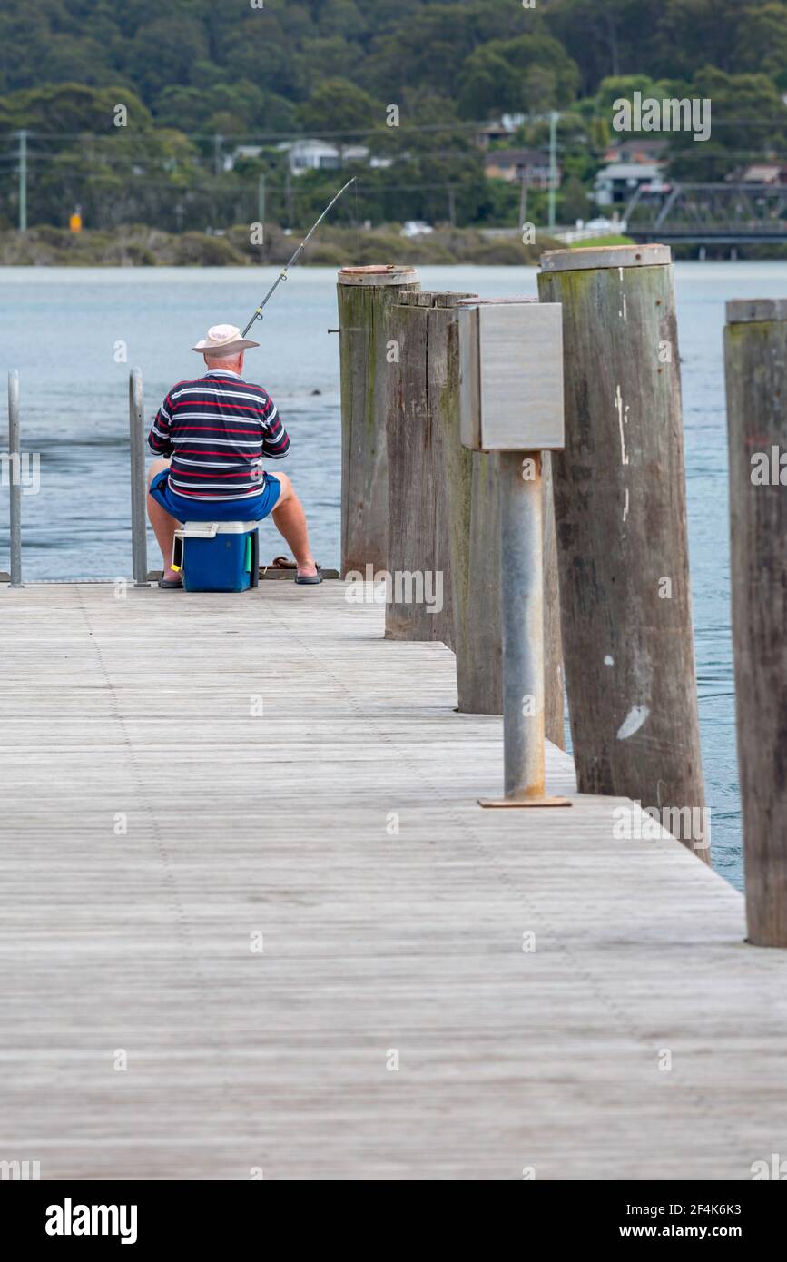 A lone older fisherman sits on an Esky cooler and fishes from a wharf