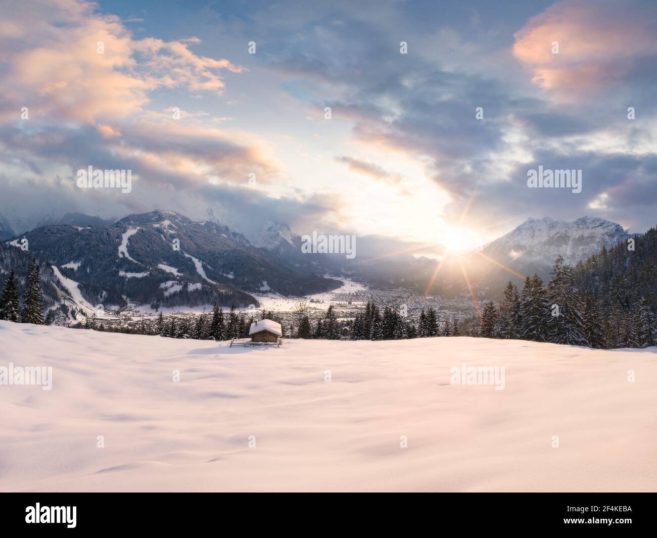 Beautiful winter scene with snow covered meadow and wooden hut in foreground - valley with Garmisch-Partenkirchen village and Alps mountains in back Stock Photo