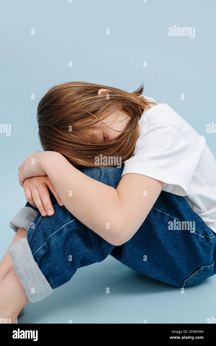 Sleepy 9 year old girl napping on her knees, on folded hands. She's sitting on the floor. Over blue background. She's wearing blue jeans and white shi Stock Photo
