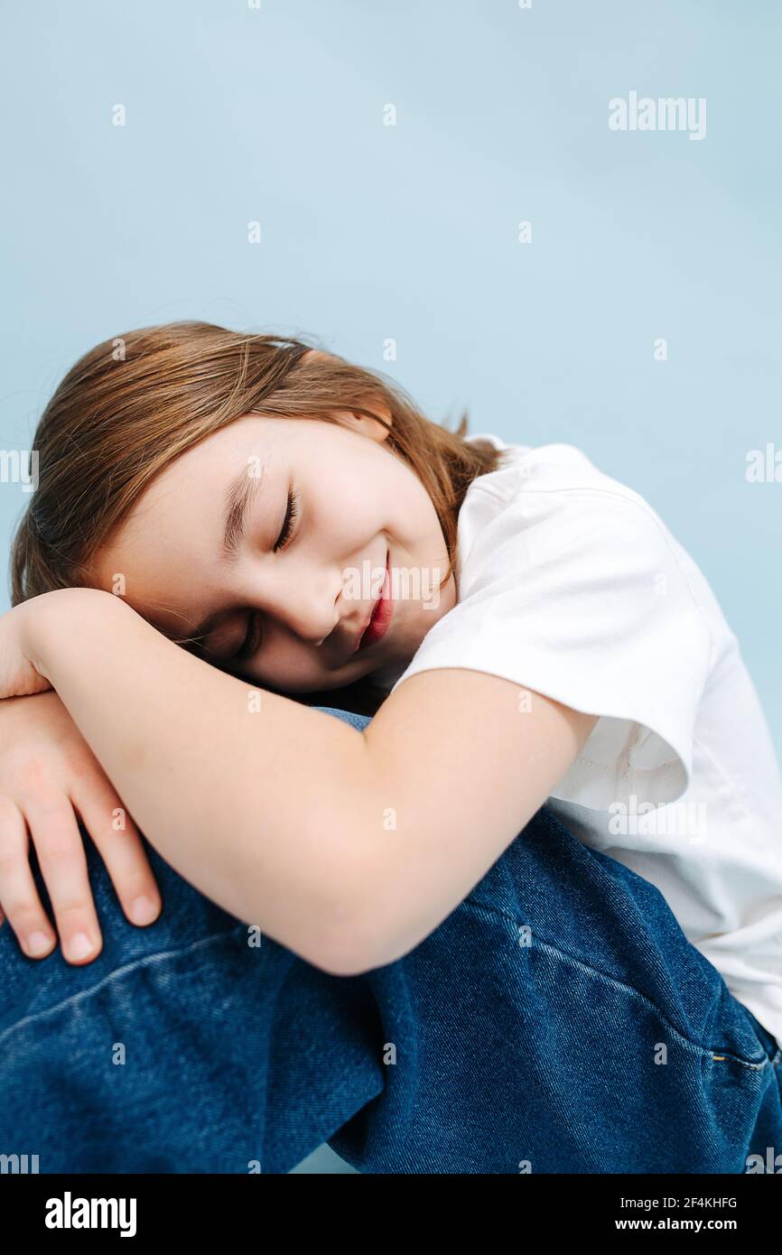 Blissful smiling 9 year old girl napping on her knees, on folded hands. She's sitting on the floor. Over blue background. She's wearing blue jeans and Stock Photo
