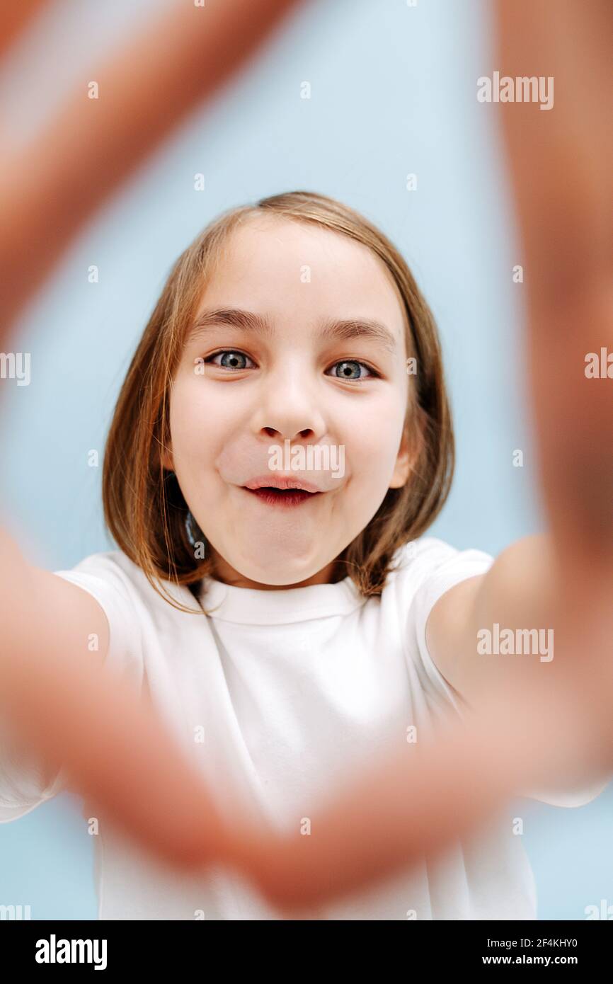 Funny smiling 9 year old girl through a finger frame she makes. Over blue background. Studio shot. She's opening her mouth, teeth covered with folded Stock Photo